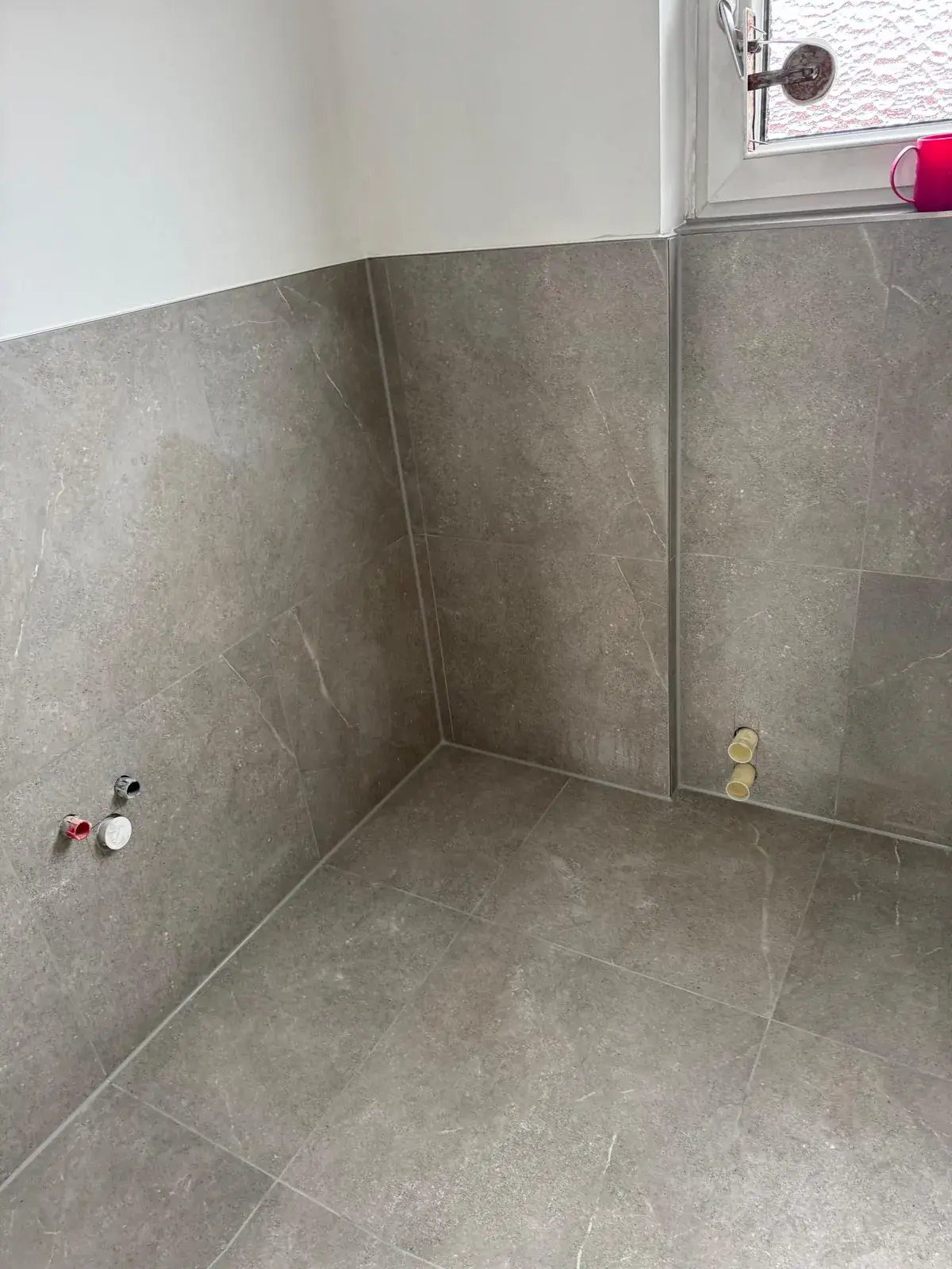 Graubraune Wand- und Bodenfliesen in einer Badecke mit vorbereiteten Wasseranschlüssen