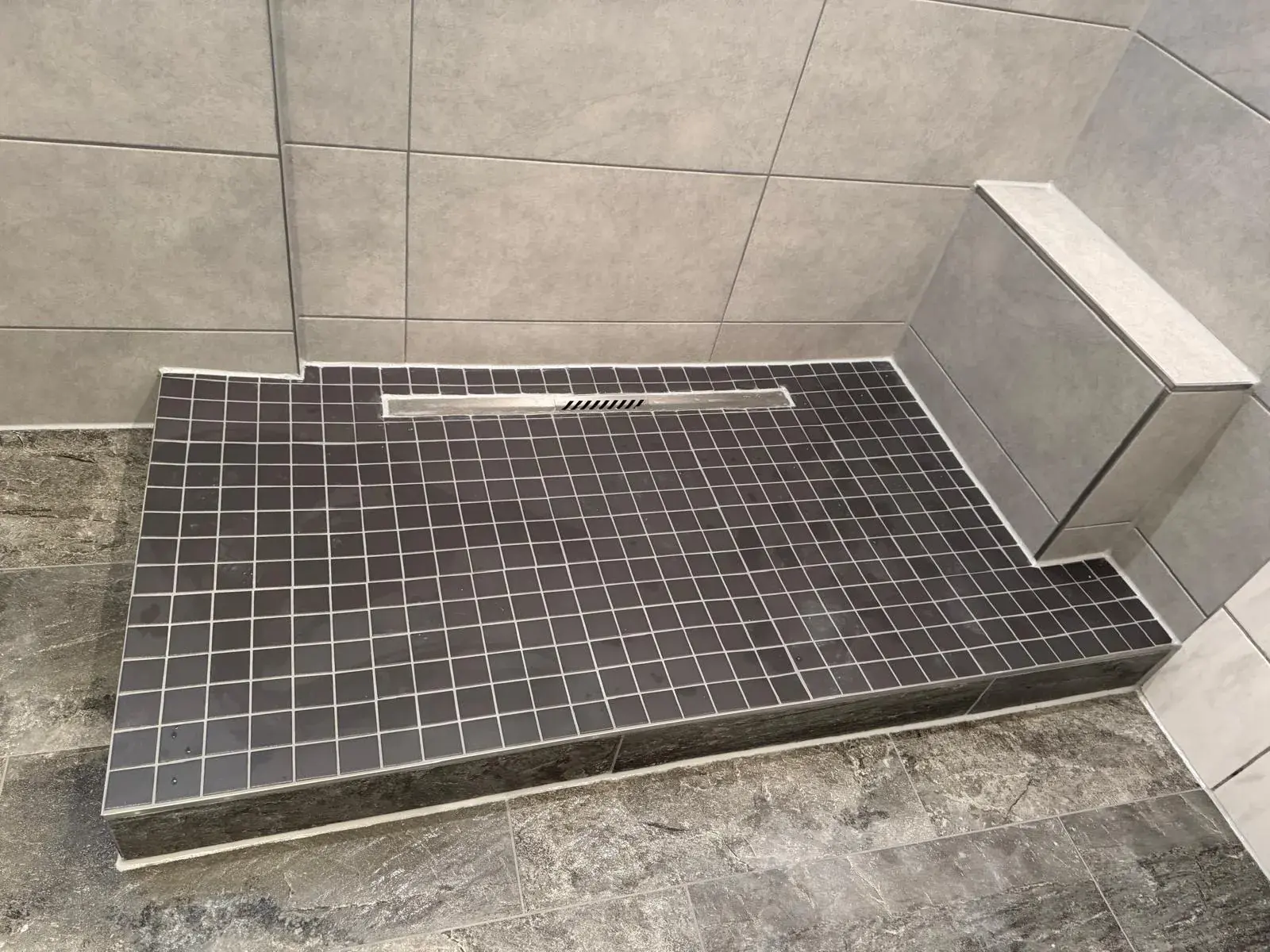 Dunkles Mosaik auf einem erhöhten Duschboden mit länglicher Duschrinne und grau gefliestem Vorsprung